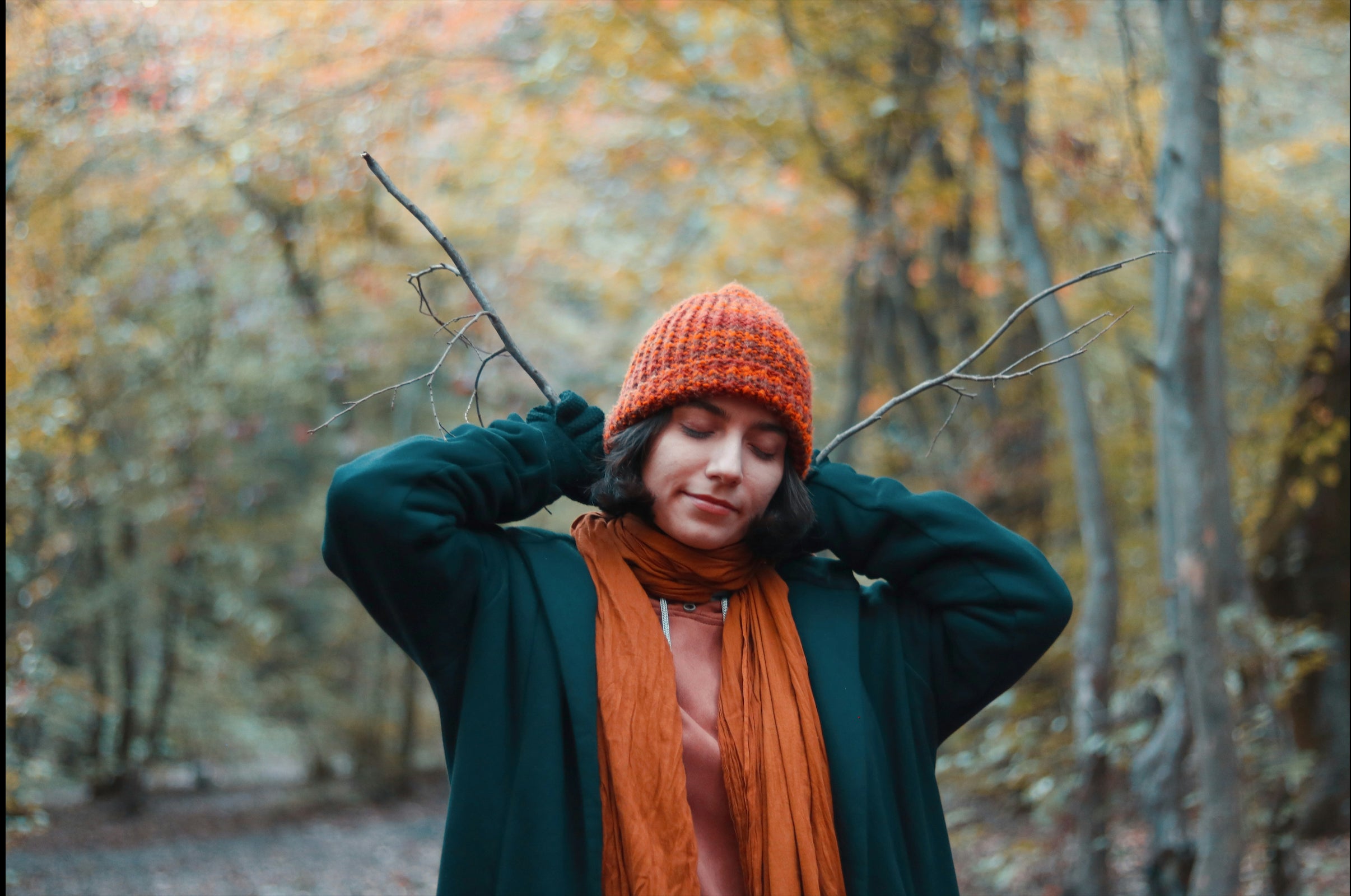 Cozy autumn style captured in a serene forest setting. A woman in WearFinds earthy-toned clothes with a knitted orange beanie, deep green oversized coat, and rust scarf, embodies warmth and comfort amid vibrant fall foliage